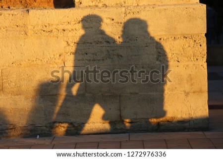 Front view of the shadows of a woman and a man walking together in the street. Figures of two persons projected at the surface of a brown textured stone wall. Silhouettes of people drawn on a facade