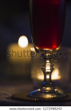 A glass of red wine on a table. In the background burning candles.