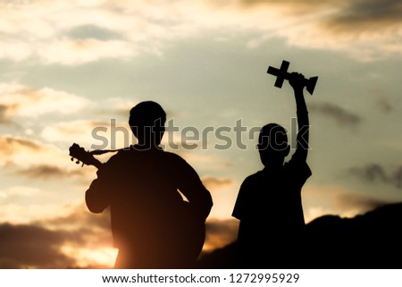 Two young christian worship God by holding cross and playing guitars with light sunset background,christian concept.