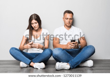 Young couple typing on phone while sitting on the floor isolated on gray background