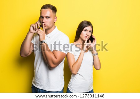 Portrait of young loving couple talking by mobile phones make silence gesture isolated over yellow background