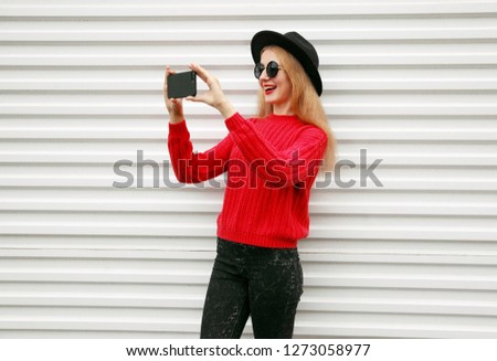 Stylish cheerful woman taking selfie picture by phone in knitted red sweater on white wall background