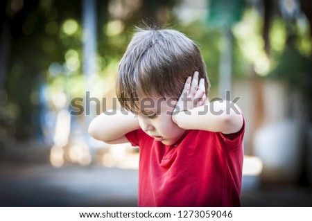 A 4 year old autistic child in a red shirt closing his ears with hands as if protecting from noise. Autism concept, Asperger syndrome, loud noise, scared little kid, parents divorce trauma. Royalty-Free Stock Photo #1273059046