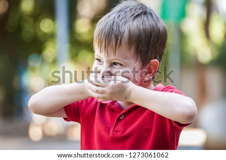 Funny Caucasian child closing his mouth to hide a smile in the park outside in the summer. Laughing positive child, happy childhood concept lifestyle, child about to laugh.