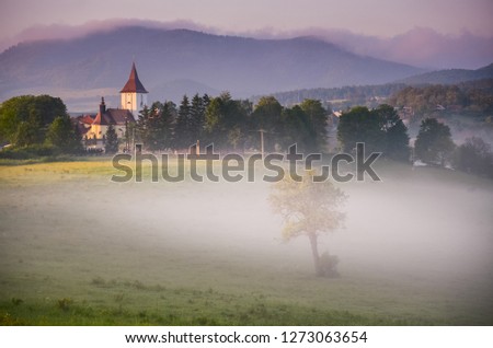 Church is hiding in the early morning fog in valley. A misty wallpaper with sunrise in the countryside.
