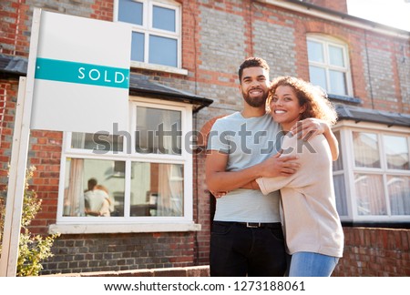 Portrait Of Excited Couple Standing Outside New Home With Sold Sign Royalty-Free Stock Photo #1273188061