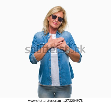 Middle age blonde woman wearing sunglasses over isolated background smiling with hands on chest with closed eyes and grateful gesture on face. Health concept.