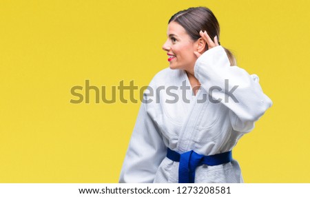 Young beautiful woman wearing karate kimono uniform over isolated background smiling with hand over ear listening an hearing to rumor or gossip. Deafness concept.