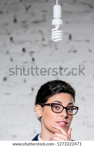 Energy efficient lightbulb hanging over young thoughtful businesswoman against brick wall at office
