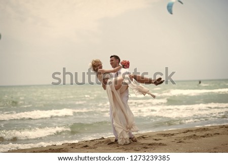 Happy beautiful smiling young wedding couple of man in white holding woman in beige dress standing on ocean beach coast on sunny weather with paraplanes on blue sky background, horizontal picture