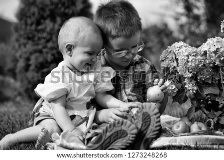Happy smiling family of red haired boy with small sister in stylish clothes sitting outdoor on picnic with basket of green apples near flowers on natural spring backdrop, horizontal picture