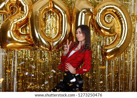 Cheerful young woman in stylish clothes posing near the background of golden balloons 2019 and showing a peace sign in the New Year's studio. Pretty girl with a beautiful smile.