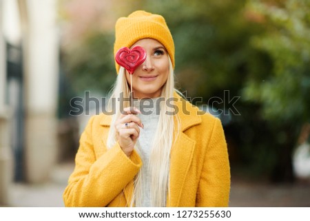Saint Valentine's day concept. Fashion portrait blond young woman in yellow coat having fun with red lollipop heart over street background. 
