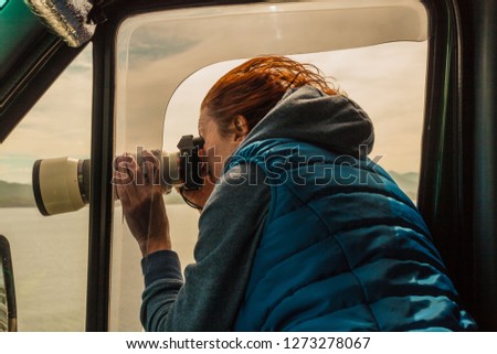 Mature woman tourist professional photographer taking photo from camper car with camera, driving on road trip. Female passenger taking picture out of window, windy weather