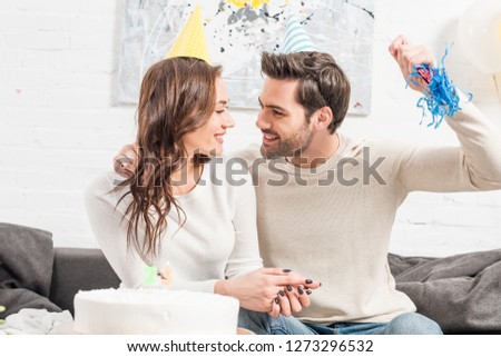 happy couple in party hats celebrating birthday with cake and party horn at home