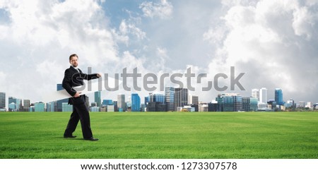 Conceptual image of young successful business man in black suit pointing aside by means of big white banner in form of arrow. Modern city buildings on background
