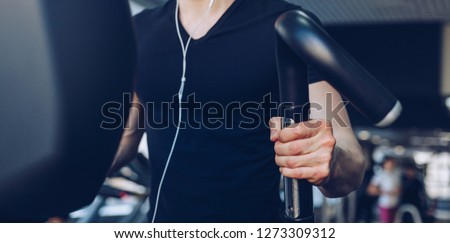 Young guy listening to music in headphones exercising in the gym on the treadmill for sports, fitness and healthy lifestyle. Hands holding the handles of the simulator close-up