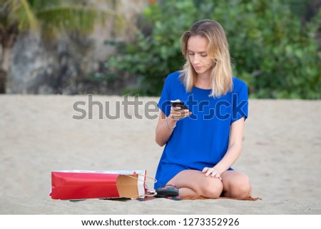A young beautiful blonde girl, on vacation at the beach, sits uses a smartphone. Portrait. 