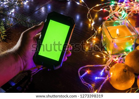 Man hand holding blank green screen mobile smart phone with abstract blurred Christmas light decorated background, home use, wooden table top view