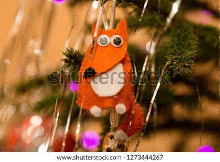 Cute handmade Christmas tree decoration in shape of nice red felted fox with big eyes. Handcrafted new year toy with background of green Christmas tree lights