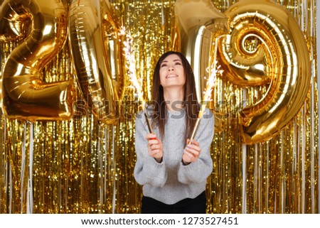 Happy joyful young woman happily holds in her hands fireworks on the background of balloons 2019 in the festive room. Cute girl celebrates the New Year. Magical celebration atmosphere.