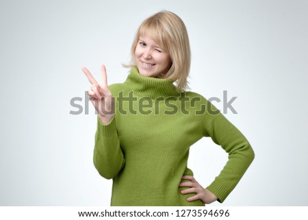 Smiling happy blonde woman in freen sweater showing victory sign and looking at camera