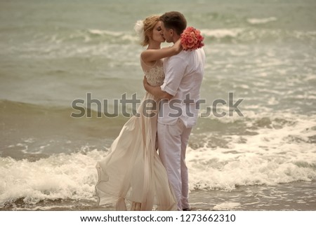 Beautiful young wedding couple of kissing man and woman in beige dress with rose bouquet emracing and standing on sea beach coast on wavy water background, horizontal picture
