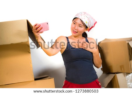young beautiful and happy Asian Korean woman excited at home living room floor unpacking belongings from cardboard boxes moving to new apartment taking selfie photo with mobile phone