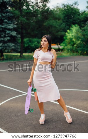 Portrait of a smiling charming brunette female holding in hand her skateboard on a basketball court. Happy woman with trendy look taking break during sunset.