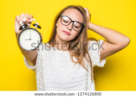 Portrait of young woman holding alarm clock looking camera isolated over yellow background