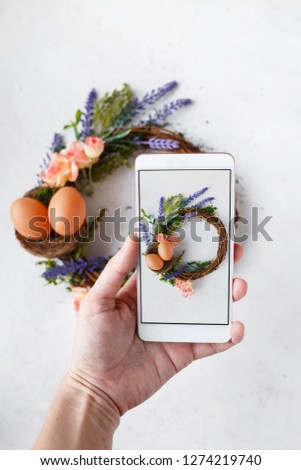 Female hand with phone photographs Easter wreath with bright spring flowers, nest with easter eggs