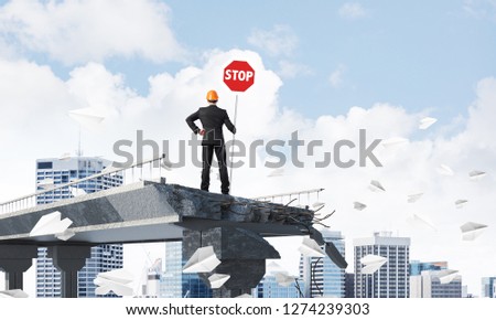 Rear view of engineer in helmet holding stop sign while standing among flying paper planes on broken bridge with cityscape on background. 3D rendering.