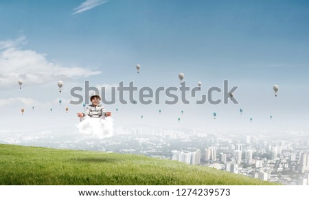 Young little boy keeping eyes closed and looking concentrated while meditating on cloud in the air with city view on background.