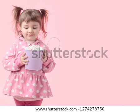 Sweet four-year-old girl in a pink jacket with white circles. Portrait of a cute little girl holding small violet bucket of marshmallow on a pink background. Copy space.