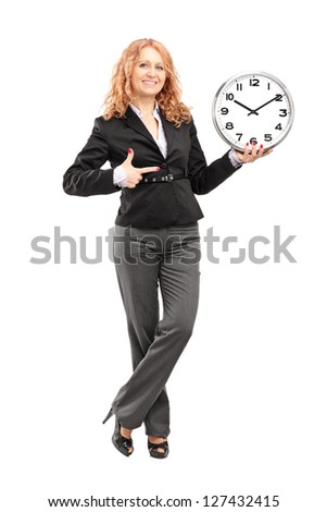Full length portrait of a blond mature woman standing and pointing on a wall clock, isolated on white background