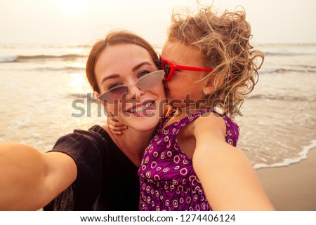 Happy stylishly mother and daughter taking selfie at sandy beach on a sunset.mothers Day.little girl blonde and beautiful woman taking pictures on the phone tourism abroad online video calling