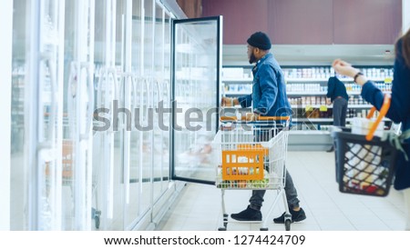 At the Supermarket: Happy Stylish Guy Pushes Shopping Cart and Chooses Frozen Vegetables in the Frozen Goods Section of the Store. Big Mall with Glass Door Fridge.