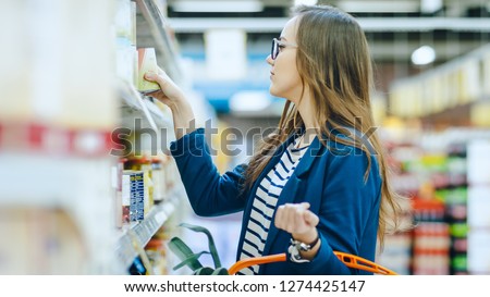 At the Supermarket: Beautiful Young Woman Browses through the Canned Goods Section of the Store. She Has Shopping Basket Full of Healthy Food Items. Royalty-Free Stock Photo #1274425147