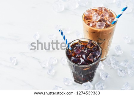 Summer drink iced coffee in a glass and ice coffee with cream in a tall glass surrounded by ice on white marble table. Selective focus, copy space for text. Top view.