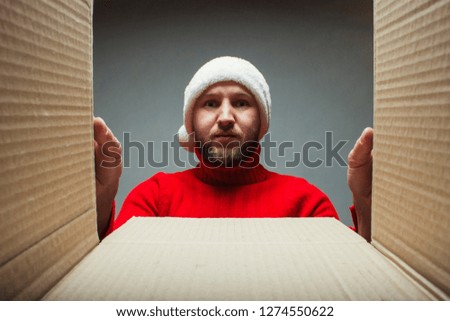 Man with a beard in a Santa Claus hat and a red sweater is looking into a box. Concept of opening gifts, surprise, pet from friends, relatives, parcel, shopping for Christmas, the new year