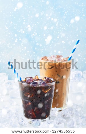 Winter drink iced coffee in a glass and ice coffee with cream in a tall glass surrounded by ice on white marble table over blue background with snow. Selective focus, copy space for text.