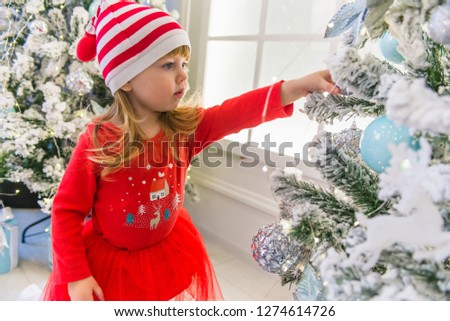 little smiling girl in a red dress and a red Christmas hat near a fireplace and a Christmas tree