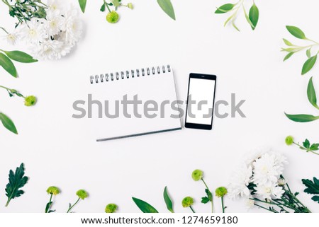 Floral flat lay arrangement of clean smart phone and empty notepad among fresh leaves and flowers, top view on white table.
