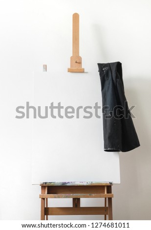 Canvas mock up with empty painting standing on wooden easel, black apron in artist studio with white wall. Interior design photography with copy space