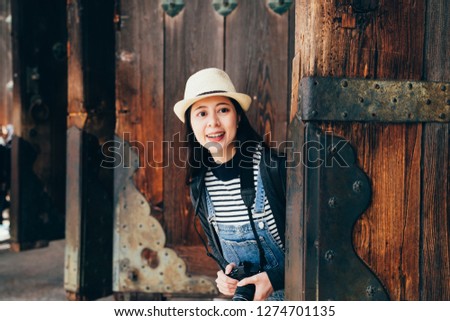curious girl traveler smiling looking from wooden door. young female asian photographer holding camera searching for amazing picture in travel trip japan. beautiful lady in straw hat cheerfully visit
