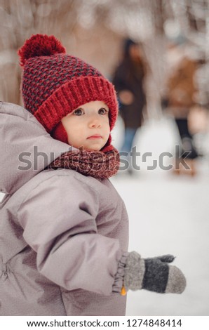 Portrait of a cute baby dressed in a gray jacket and a red hat that walks through the snow covered snow park. She smiles one in the photo during the snowfall