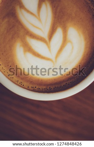 Close up white coffee cup with heart shape latte art foam on black wood table near window with light shade on tabletop at cafe