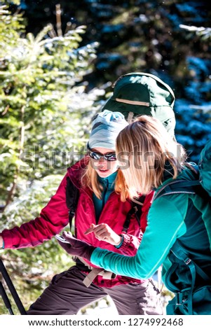Two tourists look at the phone. Two friends travel together. Women with backpacks in the winter mountains. Winter hike. Walk through the snowy forest. Mobile communication and navigation.