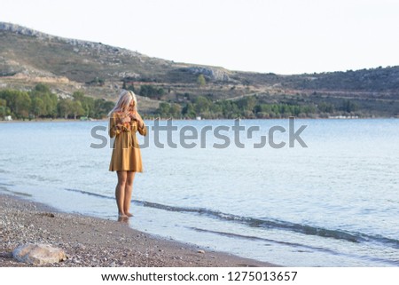 adult teenage hipster girl in yellow dress with bare feet stay in water sea shore sand coast line with mountain background scenery landscape