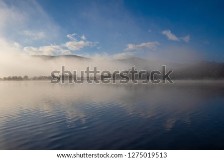 Light Fog Covers Blue Surface of Lake with Forest Background
